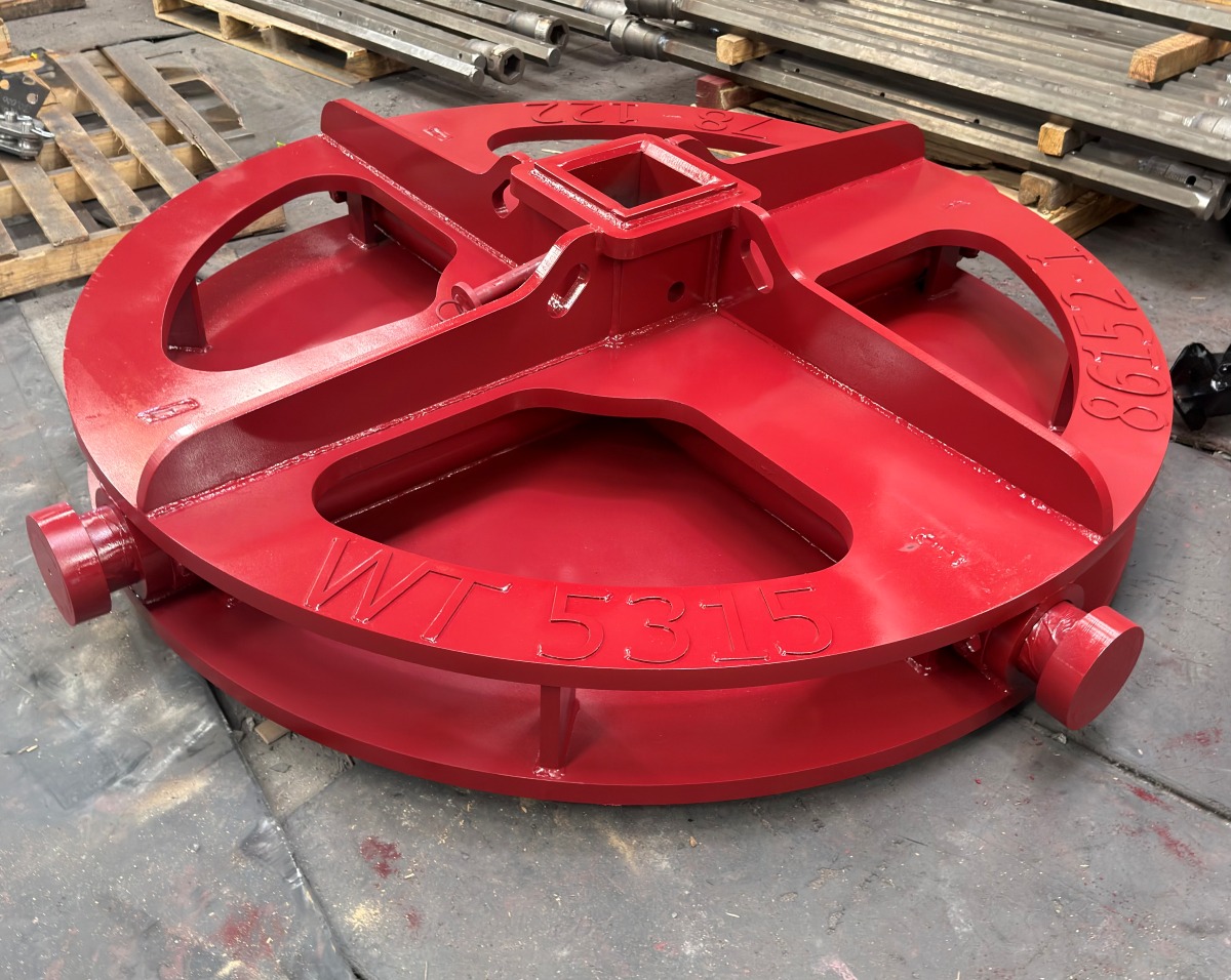 A red cylindrical metal drilling attachment on its side, resting on wooden blocks in a gravel outdoor yard.